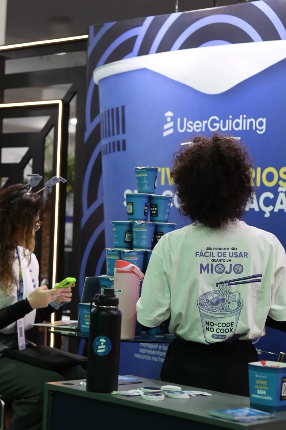 Estande da UserGuiding no Startup Summit 2026: o copo de miojo gigante com o modo de preparo, e uma visitante de camiseta no balcão